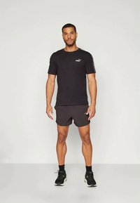 Camiseta negra de manga corta con el logo de Puma en el pecho, combinada con shorts oscuros. El modelo lleva zapatillas negras y está de pie sobre un fondo claro.