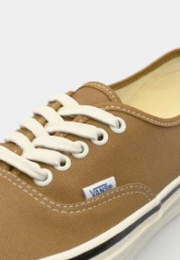 Brązowe płócienne trampki z białymi sznurowadłami i przeszyciami, z małym niebieskim logiem Vans oraz czarną gumową podeszwą. Gładka tekstura całości.