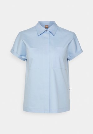 Chemise à manches courtes bleu clair avec col, deux poches poitrine et une petite étiquette de marque au niveau de l'encolure.