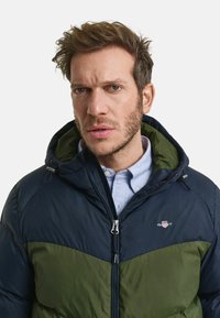 Marineblau und olivgrüne Steppjacke mit Reißverschluss, Kapuze und Logodetails. Strukturiertes Gewebe, gesteppte Optik und kontrastierende Farbbereiche.