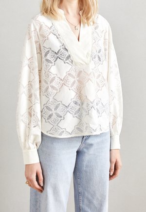 Vrouw draagt een witte blouse met lange mouwen, een bloemig kanten patroon en een V-hals, gecombineerd met lichtblauwe spijkerbroek.
