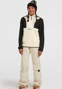 Kremowo-czarna polarowa bluza z kołnierzem na zamek, zestawiona z beżowymi śniegowcami i brązowymi butami, noszona z czarną dzianinową czapką.
