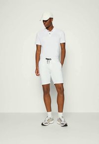 Weißes Poloshirt, weiße Shorts mit Kordelzug, weiße Mütze, graue und silberne Sportschuhe, stehend vor einem schlichten Hintergrund.