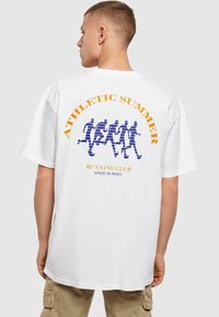 Hvid bomulds t-shirt med en afslappet pasform. Har et blåt grafisk motiv af løbende figurer og orange tekst, der siger "ATHLETIC SUMMER RUNNING CLUB."