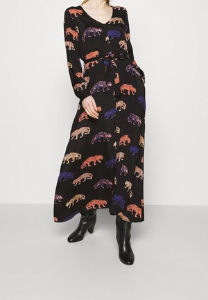 Femme portant une robe noire à manches longues boutonnée avec des imprimés léopard multicolores et des bottines noires, debout avec une main dans la poche.
