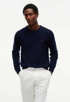 Man wearing a navy blue cable knit sweater and white pants with hands in pockets, standing against a plain white background.