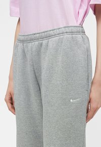 Άτομο που φοράει ανοιχτό γκρι φόρμα Nike και ανοιχτό ροζ μπλουζάκι, που δείχνει ελαστική μέση και το λογότυπο Nike στον αριστερό μηρό.