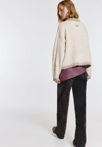 Cremefarbener gestrickter Pullover mit hohem Kragen, kombiniert mit dunkler, strukturierter Hose. Auffällige violette Lederakzente sind am Bund sichtbar.