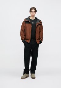 Καφέ αδιάβροχο μπουφάν με μαύρες λεπτομέρειες, μαύρο μπλουζάκι, μαύρο παντελόνι cargo και γκρι μπότες. Απλός σχεδιασμός, ελάχιστες λεπτομέρειες.