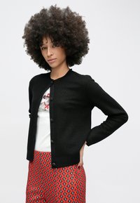 Kährikutega naine musta nööpidega kardiganiga, valge looma mustriga särgiga ja punaste mustriliste pükstega, poseerib käsi puusal.