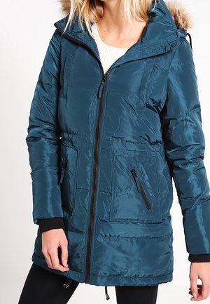 Frau trägt einen türkisfarbenen, gesteppte Wintermantel mit pelzbesetzter Kapuze, Frontreißverschluss und Reißverschlusstaschen über einem weißen Pullover.