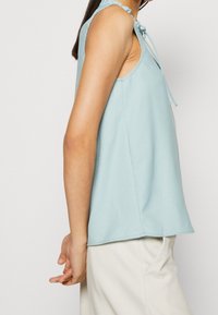 Top sin mangas de color azul claro con un corte holgado, que cuenta con un cuello con volantes y un detalle de lazo, hecho de una tela suave. No se observan patrones ni herrajes.