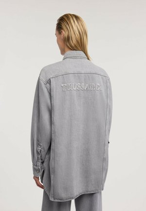 Blonde Person trägt eine lockere graue Jeansjacke mit "TRUSSARDI" auf dem oberen Rücken bestickt, steht vor einem einfarbigen hellen Hintergrund.