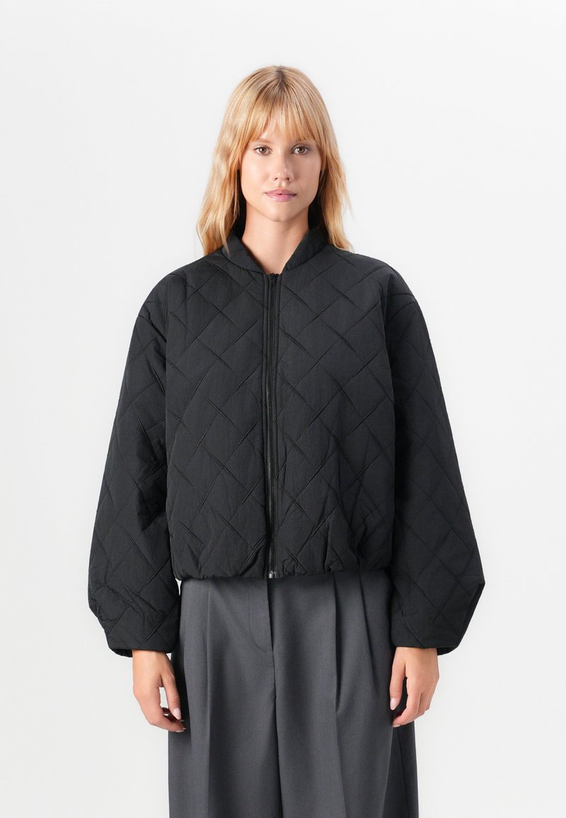 Femme portant une veste noire matelassée zippée avec des manches bouffantes et un pantalon large plissé gris, se tenant devant un fond blanc.