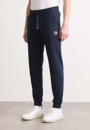 Pantaloni della tuta blu navy con fascia elastica in vita e coulisse, dotati di tasche laterali e toppa con logo rosso e bianco. Abbinati a sneakers bianche.