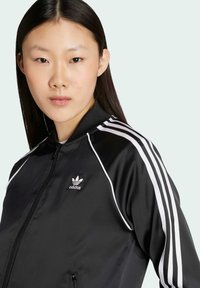 Crna sportska zip jakna izrađena od glatkog materijala, s bijelim prugama na rukavima i malim bijelim Adidas logotipom na prsima.