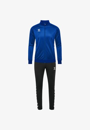 Blaue Zip-Jacke mit hohem Kragen, kombiniert mit schwarzen Jogginghosen mit weißen Streifen und Logodetails. Glattes, sportliches Material.