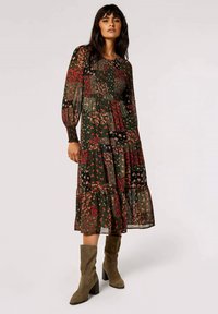 Kleid mit floralem Muster in Dunkelgrün, mit langen, durchsichtigen Ärmeln, geschmücktem Oberteil und gestuftem Volantabschluss. Kombiniert mit olivgrünen, kniehohen Stiefeln.