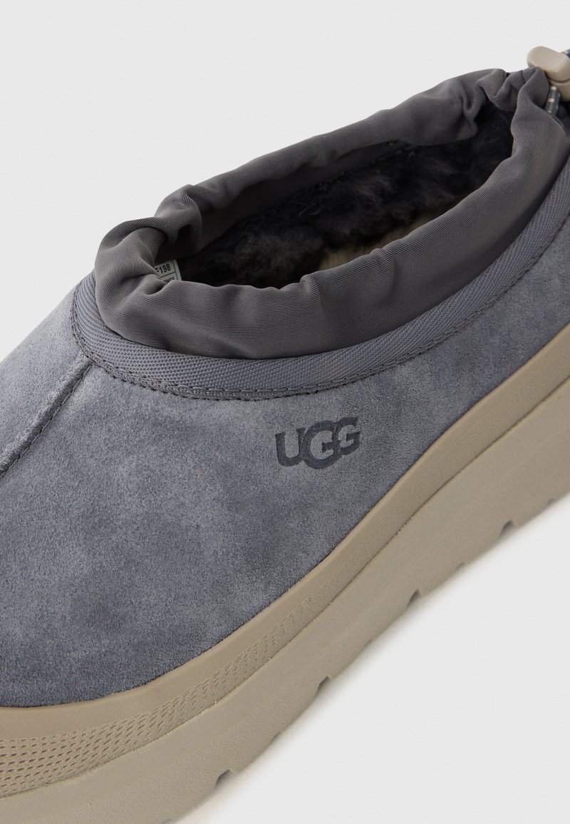 Chaussure slip-on en suède gris avec une doublure intérieure douce, un col élastique et une semelle en caoutchouc beige. Présente le logo UGG sur le côté.