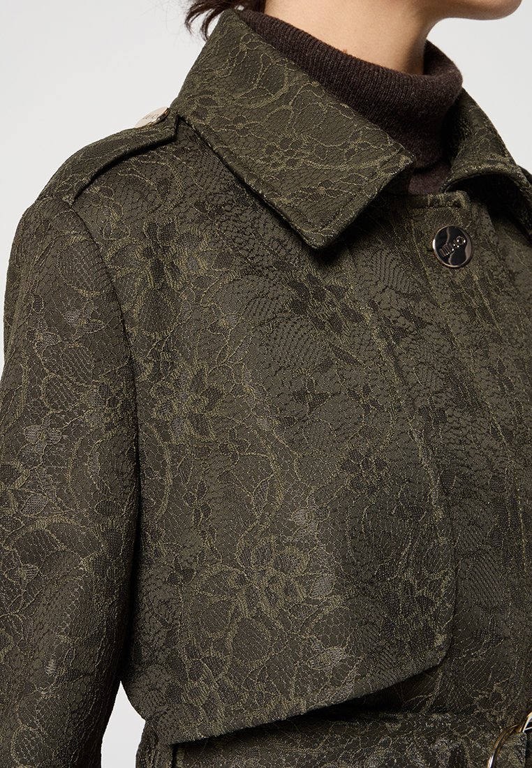 Giacca verde oliva con texture in pizzo floreale, colletto abbottonato, spallina con bottone, indossata sopra un maglione a collo alto marrone scuro.
