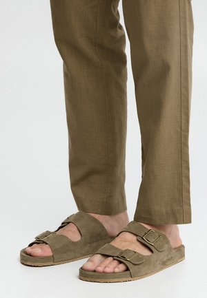 Legs wearing light brown trousers and matching suede sandals with two adjustable buckled straps on each foot.