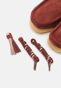 Chaussons en daim marron avec une semelle en tan douce, accompagnés de trois glands marron et beige et de cordons pour la personnalisation.
