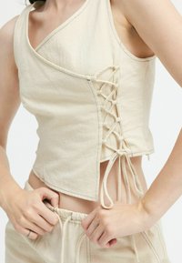 Beige Cropped-Top mit asymmetrischem Ausschnitt und seitlichem Schnürdetail. Aus strukturiertem Stoff mit sichtbaren Nähten entlang der Kanten gefertigt.