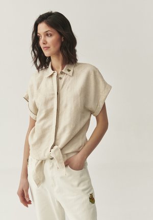 Beige linnen overhemd met korte mouwen en een strik in de taille, voorzien van een subtiele bloemenpatroon en een decoratieve speld op de kraag. Witte broek met borduursel.