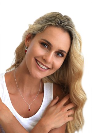 Collier en argent avec un pendentif en goutte rouge, accompagné de boucles d'oreilles rouges assorties. Le modèle porte une chemise blanche et sourit.
