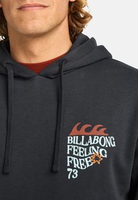 Close-up of a person wearing a dark hoodie with a Billabong logo featuring waves, the phrase "Feeling Free," and the number 73.