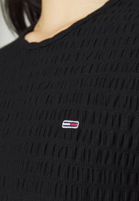 Black textured fabric top with a small rectangular logo in white, navy, and red on the chest near a woman's neck and shoulder.