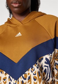 Mikina s kapucí, horní část v hnědé barvě s modrým šípovým vzorem a spodní část s leopardím potiskem v béžových a modrých tónech.