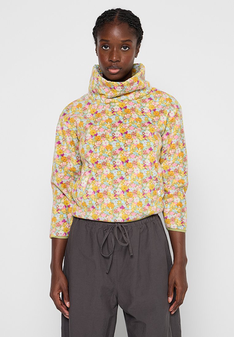 Vrouw in een pastelgekleurde, gebloemde fleece pullover met een hoge kraag en grijze joggingbroek, staande tegen een eenvoudige witte achtergrond.
