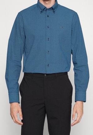 Homme portant une chemise bleue à motifs boutonnée, rentrée dans un pantalon de costume noir, les mains détendues le long du corps.