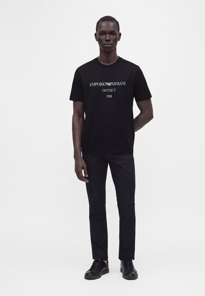 Άνδρας με ολομαύρο ντύσιμο, πουκάμισο Emporio Armani μαύρο, μαύρο παντελόνι και μαύρα αθλητικά παπούτσια, στέκεται μπροστά σε απλό λευκό φόντο.