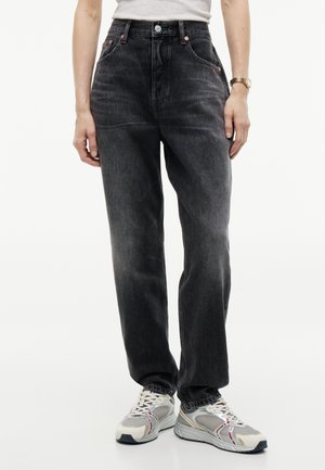 Personne portant un jean noir taille haute et des baskets blanches de sport, debout devant un fond blanc uni.