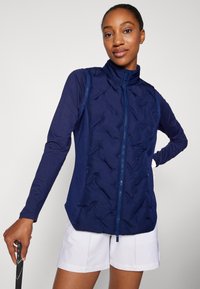 Tamno plavi prsluk s kvadratnim dizajnom, visokim ovratnikom i punim zatvaračem. Nosivi preko majice s dugim rukavima, u kombinaciji s bijelim šortsem, drži u ruci tenis racket.