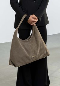 Suède handtas in taupe met een slouchy vorm, een lange schouderband en een goudkleurige logo plaat. Gladde textuur met minimale hardwaredetails.