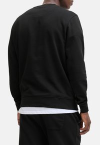 Black sweatshirt featuring a round neckline, ribbed cuffs, and hem, with a smooth texture. Worn over a white layered shirt.