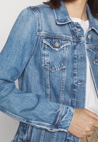 Jeansjacka i ljusblått med klassisk krage, två bröstfickor, silverknappar med tryck och lätt sliten textur.