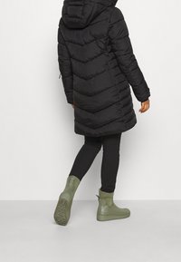 Abrigo acolchado negro con capucha, con un diseño de acolchado en forma de chevrón, combinado con botas de goma verdes hasta el tobillo. La modelo lleva leggings negros.