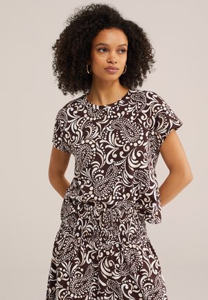 Ženska z kodrastimi lasmi, oblečena v ujemajočo se kratko majico z vzorcem v temno rjavi in beli barvi ter krilo, stoji z rokami za hrbtom.