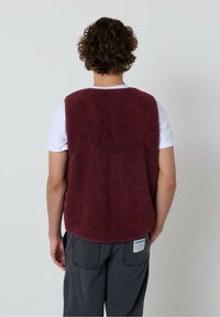 Cilvēks ar cirtainiem matiem, ģērbies baltā īsām piedurknēm kreklā un bordo krāsas vilnas veste, stāv pretī vienkāršai gaiši pelēkai siena.