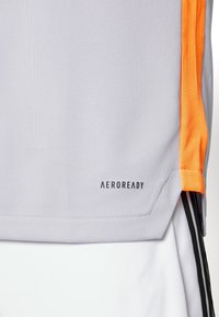 Camiseta deportiva de color gris claro con acentos de malla naranja, textura suave y un logo de "AEROREADY" cerca del dobladillo. Presenta rayas negras en los laterales.