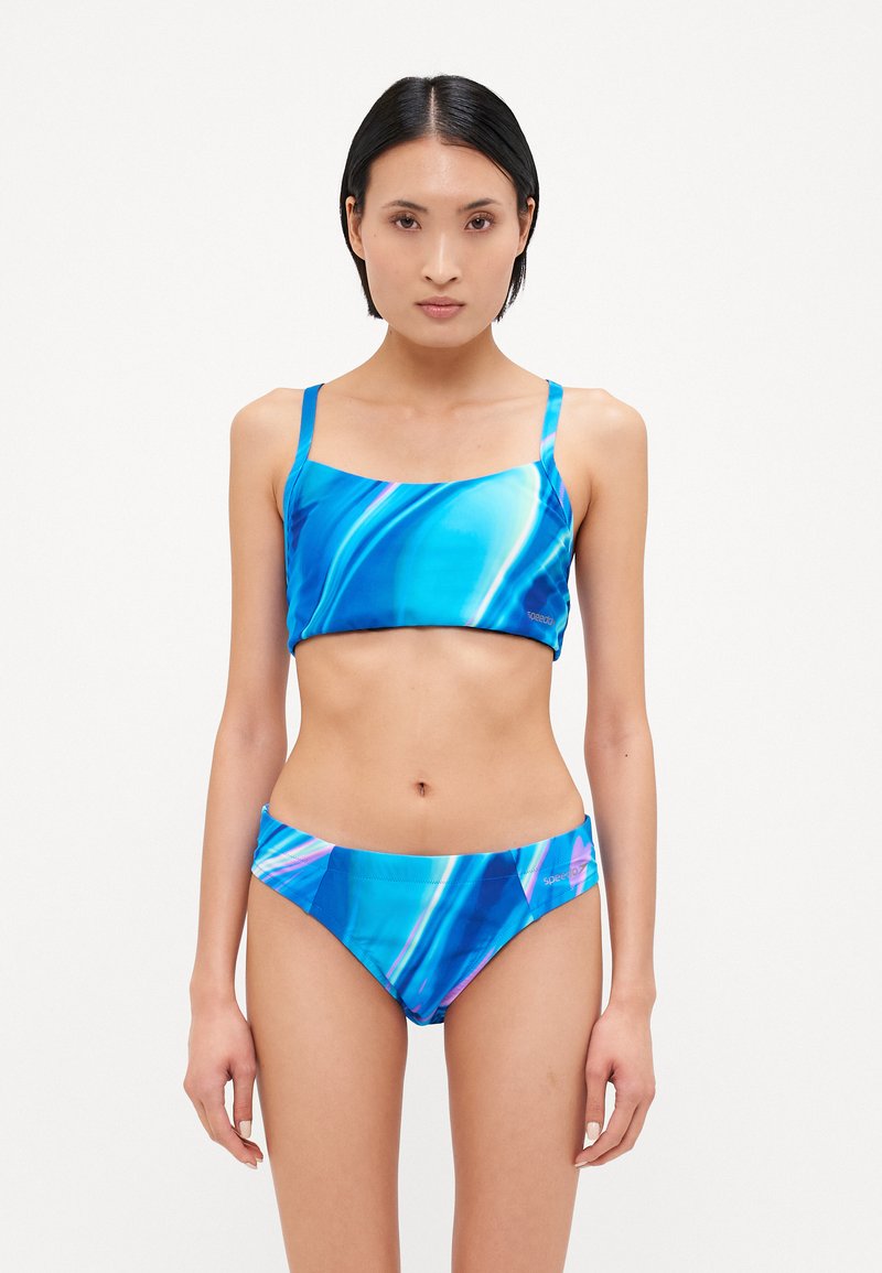 Modrý športový bikini set obsahujúci top so štvorcovým výstrihom na tenké ramienka a zodpovedajúce bikini spodky, oba so vzorom vlny v rôznych odtieňoch modrej.