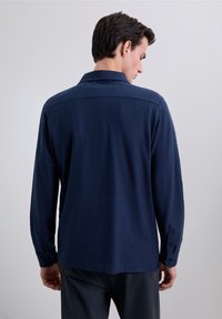 Camisa de manga larga azul marino con cuello abotonado, textura suave y aberturas laterales. Presenta puños de dos botones y una sisa trasera para mayor estructura.