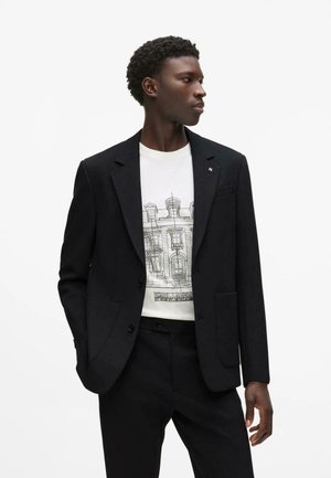 Muž v čiernom obleku a bielom tričku s architektonickým náčrtom, stojí s jednou rukou v vrecku, pozerá sa na stranu.