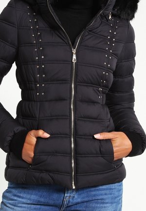 Schwarze gesteppte Jacke mit vorderem Reißverschluss, Nietenverzierung, Seitentaschen und gerippten Bündchen, getragen mit blauer Jeans.