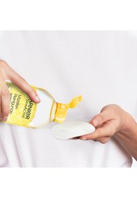 Garnier micellär rengöringsvatten i en klar flaska med ett gult fliplock, som hälls på en rund, texturerad bomullsrondell som hålls av en hand.
