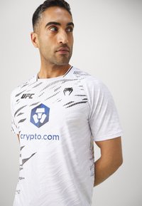 Camisa atlética blanca con patrones abstractos negros, de mangas cortas y cuello redondo. Presenta los logotipos de UFC y crypto.com exhibidos de manera prominente.